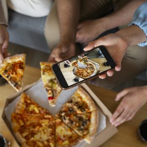 Friends sitting on the couch, taking a photo of their pizza with a cell phone to share on social media.