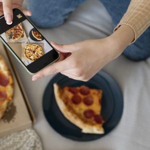 A woman kneels beside a pepperoni pizza. She is taking a photo with her cell phone to share on social media.